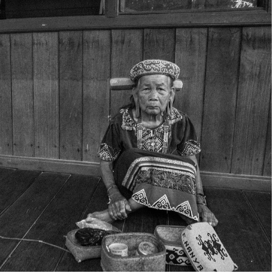 An elderly woman in traditional headdress, seated with her wares on a wooden floor, from REINKARNASI