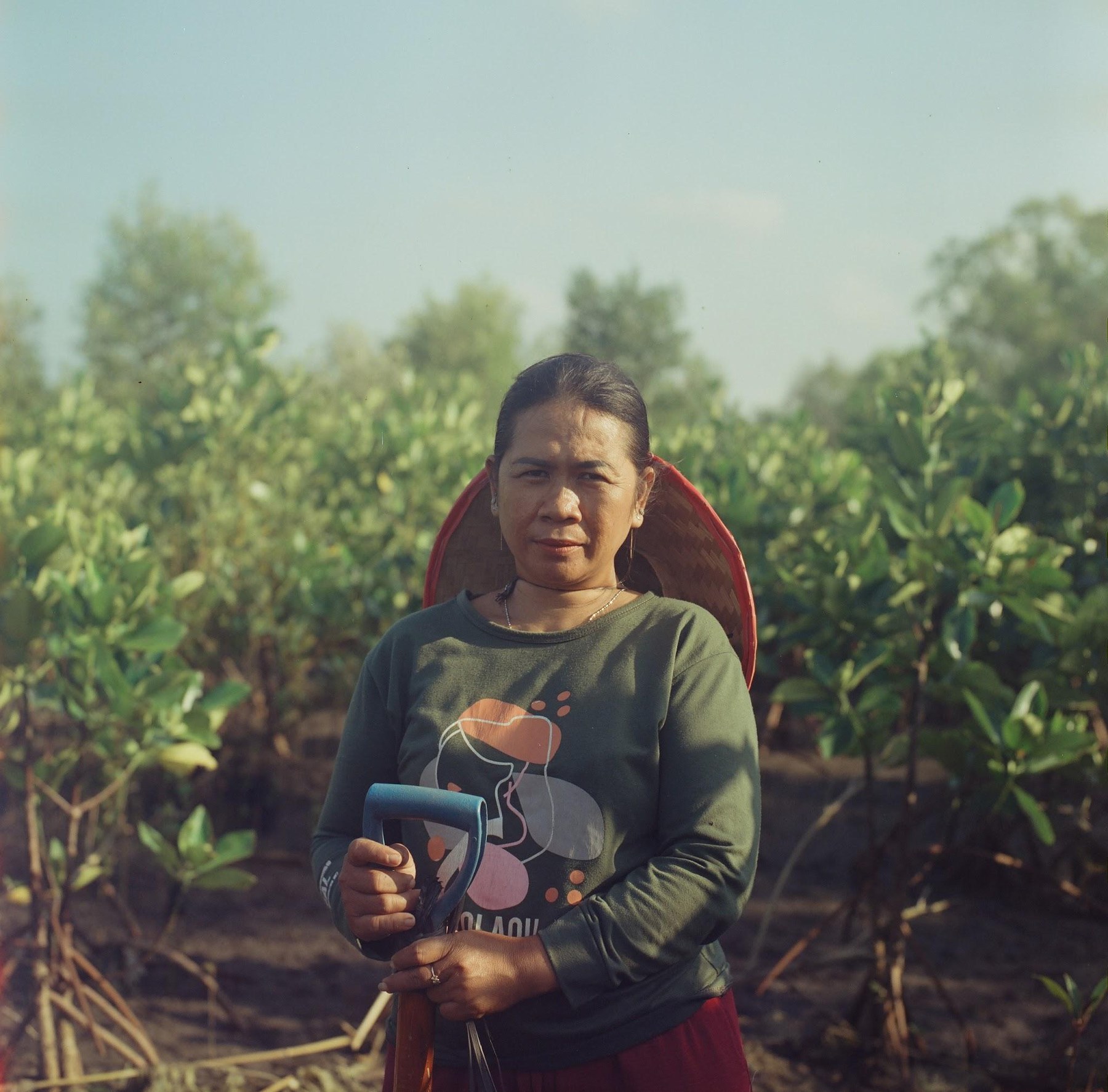 Portrait of a woman in a green shirt holding a shovel, mangrove trees behind her