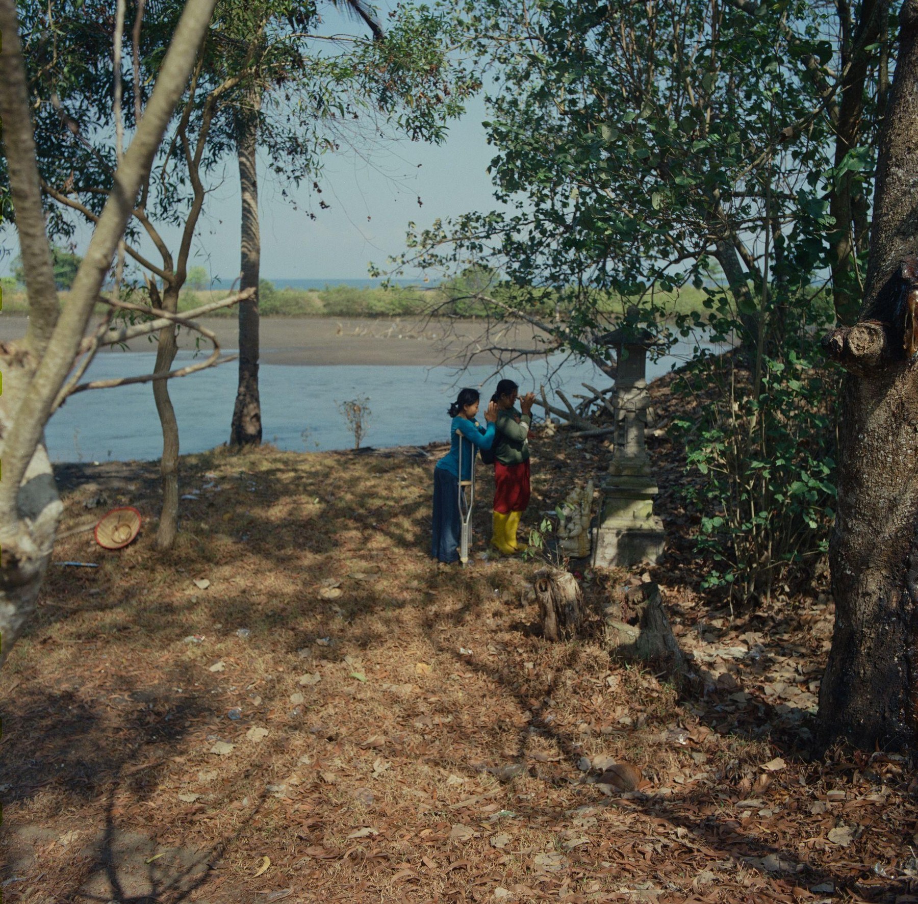 Two women praying at a small shrine under the trees by the water
