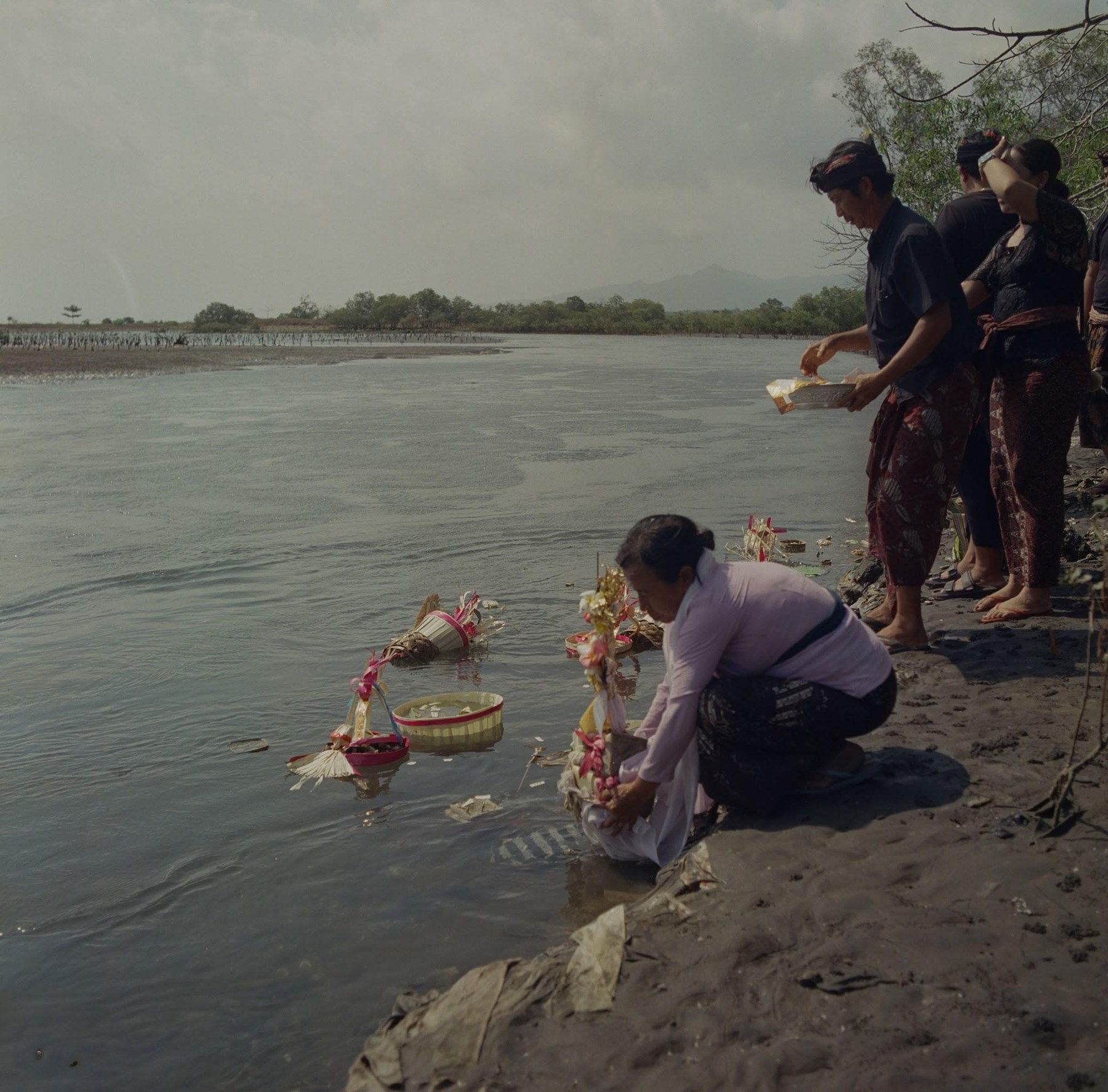 A woman kneeling at the water to release ashes and offerings into the sea