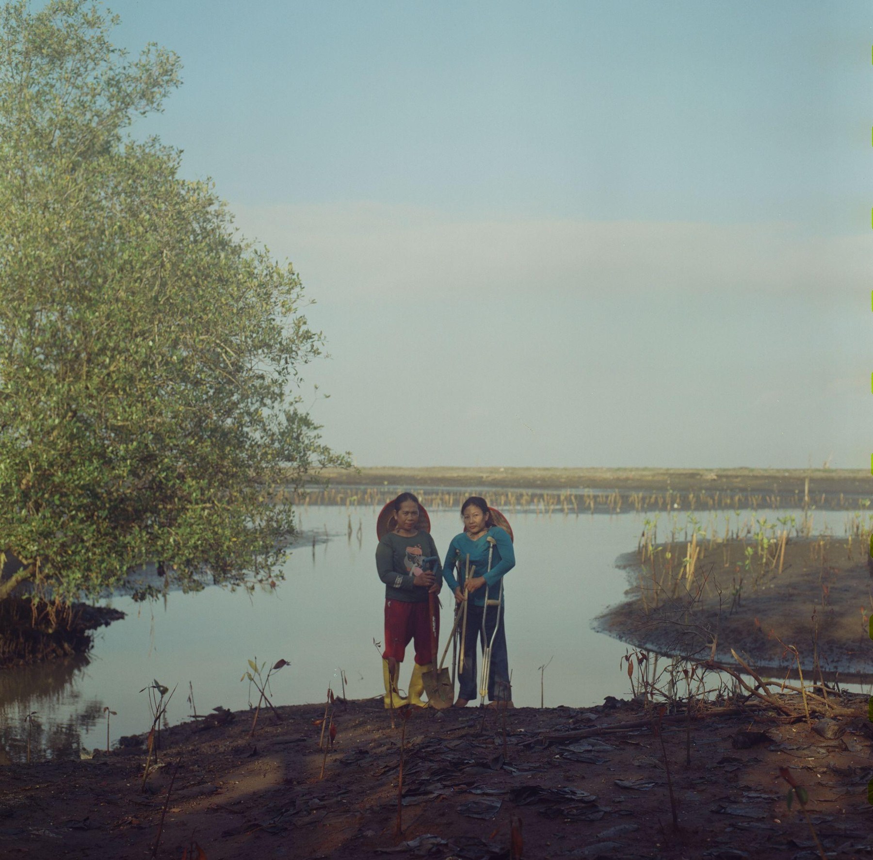 Two women standing on a mud bank at the edge of the water, mangroves beside them