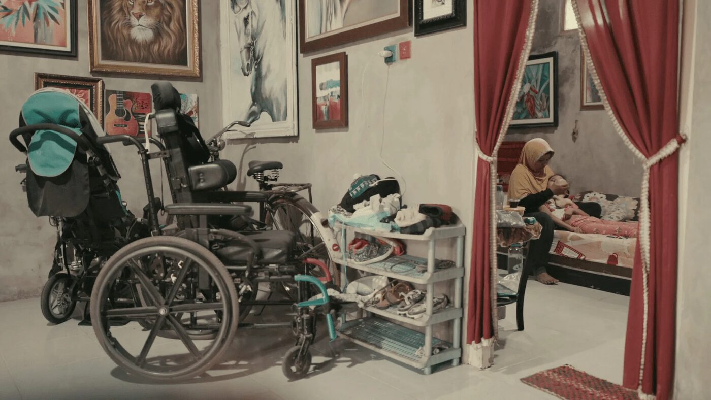 Wheelchairs and mobility aids in a family home, a mother tending to her daughter on a bed beyond the doorway, from Atas Nama Daun