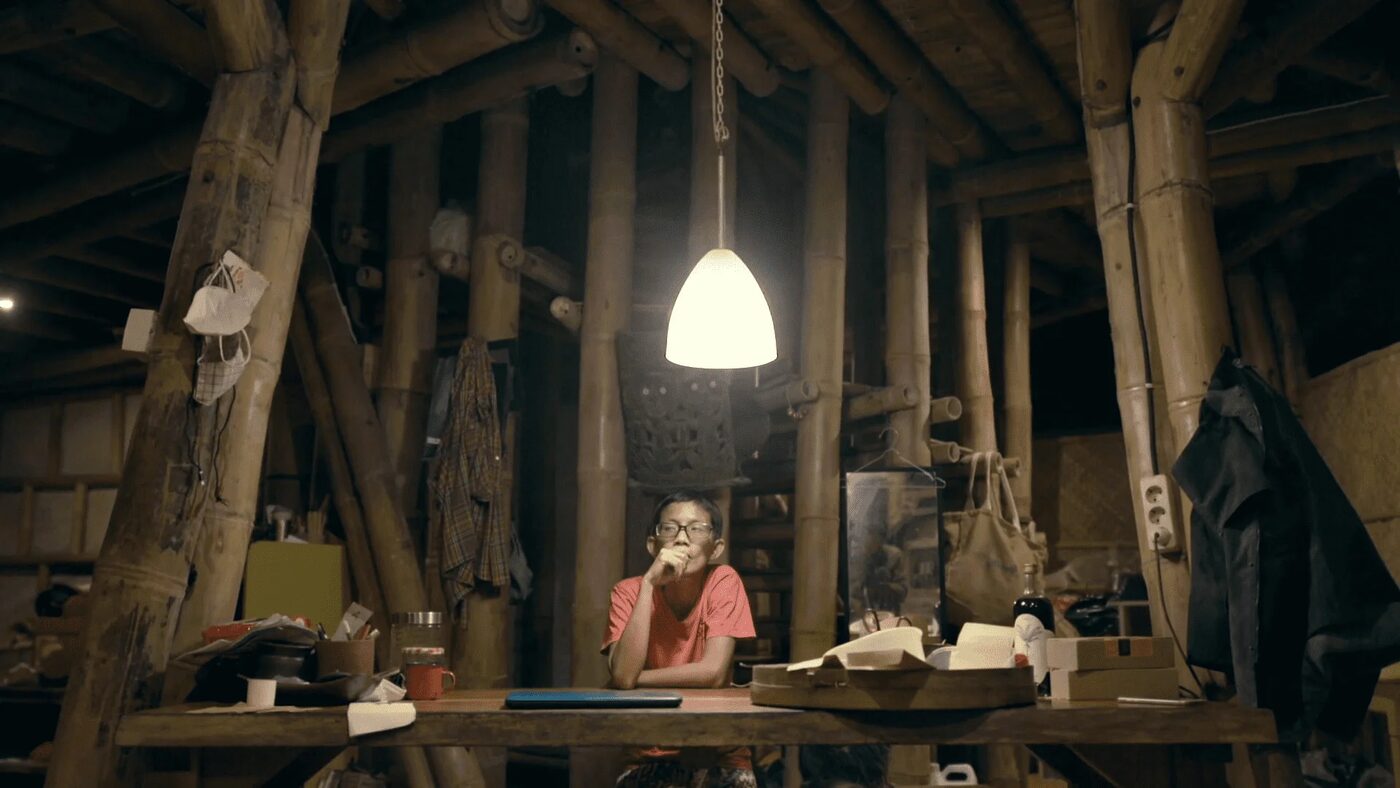 A man sitting alone at a table under a hanging lamp in a bamboo house at night, from Atas Nama Daun