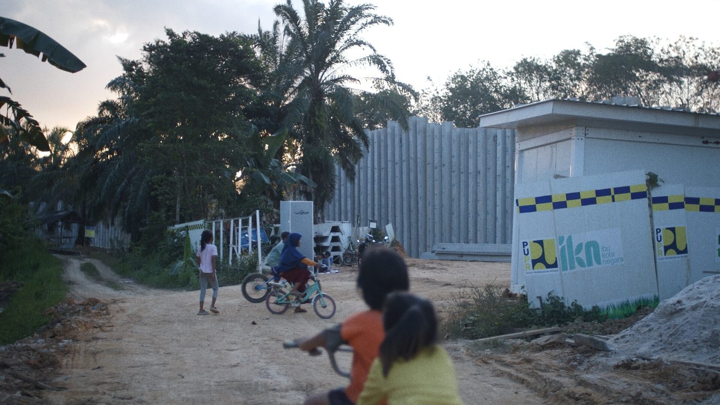 Children cycling past a construction hoarding for Indonesia’s new capital, from A Tale of the Stolen River